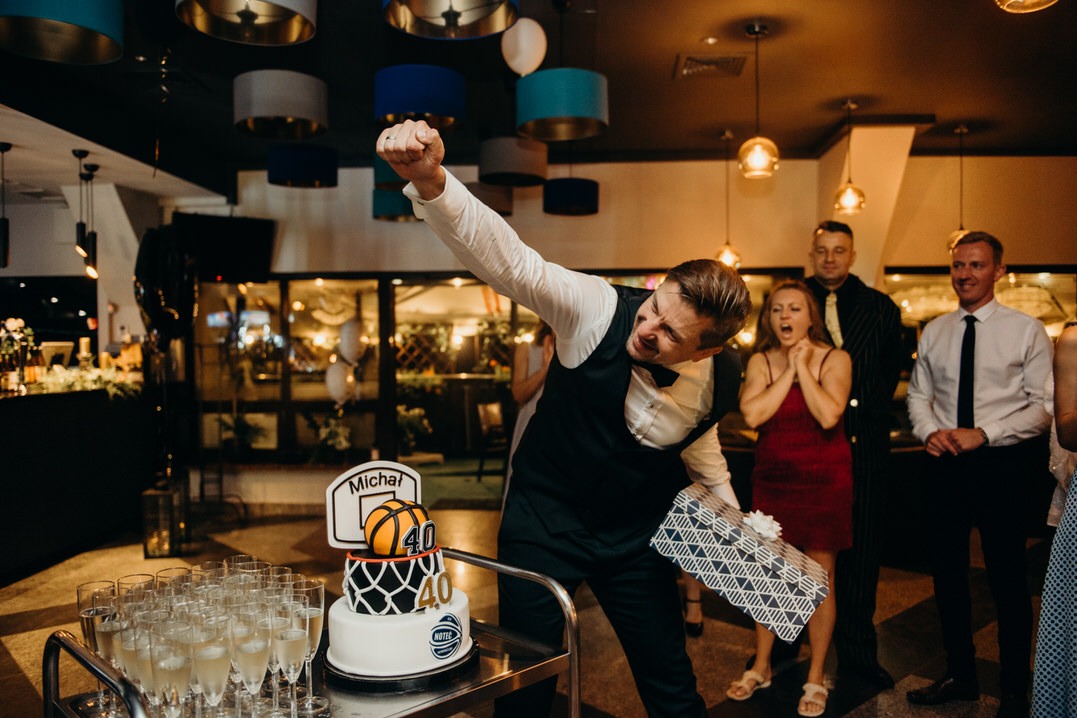 Uczestnik imprezy wznosi pięść w geście triumfu obok tortu urodzinowego z motywem koszykówki i napisem '40', podczas reportażu z 40-tych urodzin, w tle rozemocjonowana kobieta i obserwujący goście.
