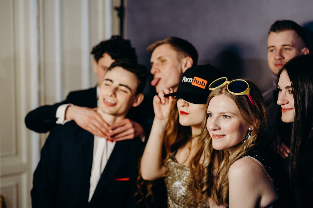 Grupa młodych ludzi pozuje do zdjęcia w fotobudce, w tle ściana z beżowymi panelami, jedna z osób ma na sobie czapkę z logotypem.