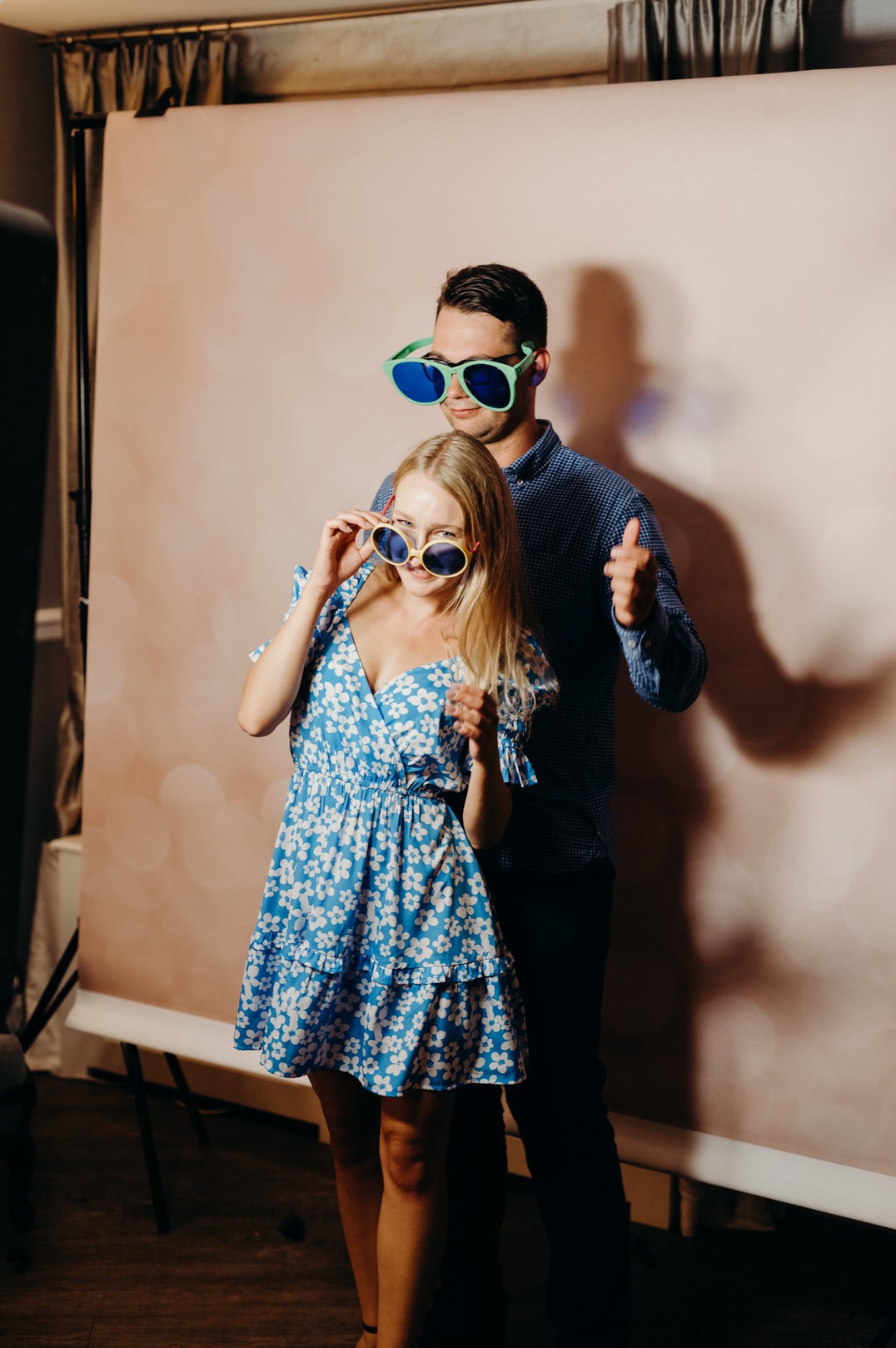 Para w zabawnych, przerysowanych okularach pozuje przed tłem fotobudki, kobieta w kwiecistej sukience uśmiecha się, mężczyzna pokazuje kciuk w górę.