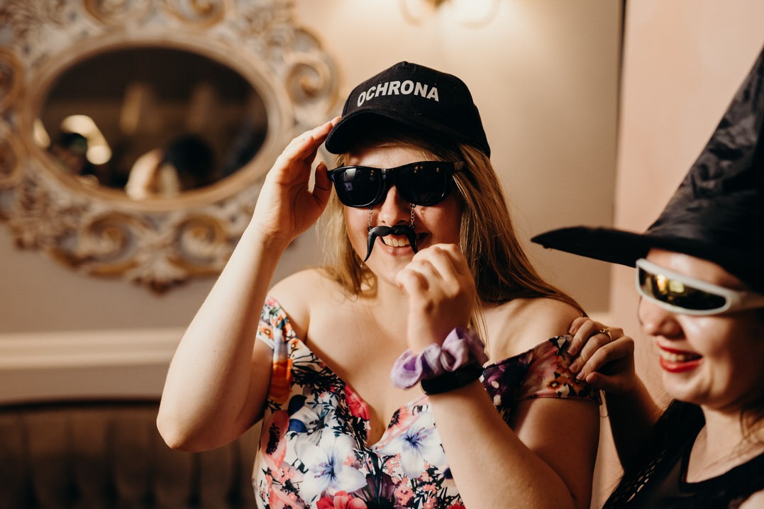 Dwie kobiety w przebraniach w fotobudce: jedna w czapce z napisem 'OCHRONA' i sztucznym wąsem na łańcuszku, druga w czarnym kapeluszu czarownicy i futurystycznych okularach.