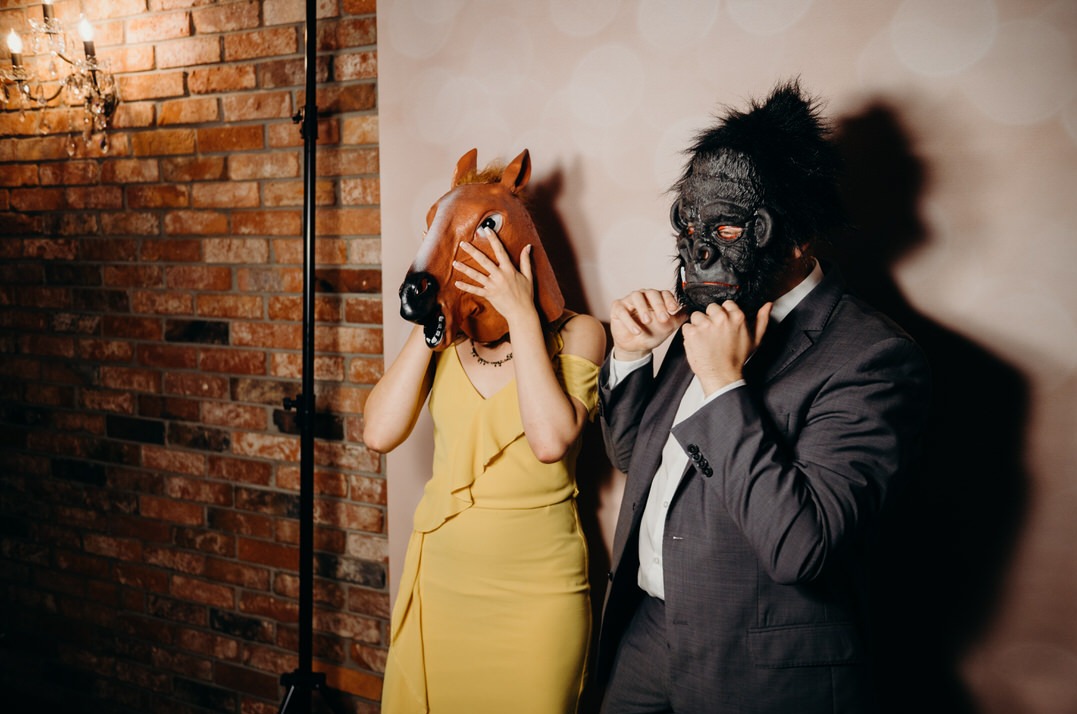 Para w maskach konia i goryla pozuje do zdjęcia przy ceglanej ścianie, mężczyzna w garniturze, kobieta w żółtej sukience.