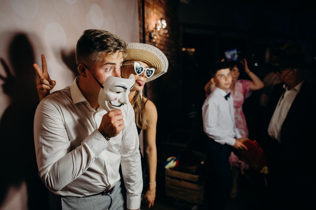 Grupa osób pozuje z rekwizytami: mężczyzna trzyma maskę uśmiechniętej twarzy, kobieta w kapeluszu i okularach w kształcie serc wystawia język, w tle inni goście.