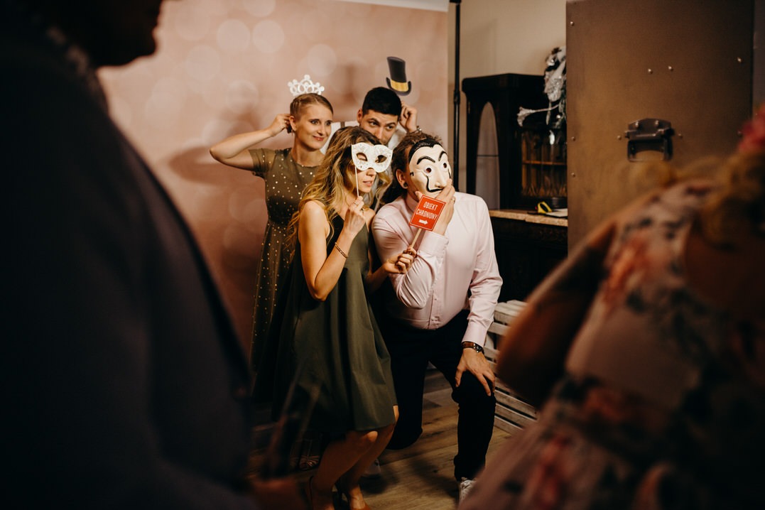 Grupa czterech osób pozuje do zdjęcia w fotobudce, trzymając maski i rekwizyty na patyku, w tle dekoracyjna ściana i ciemne meble.