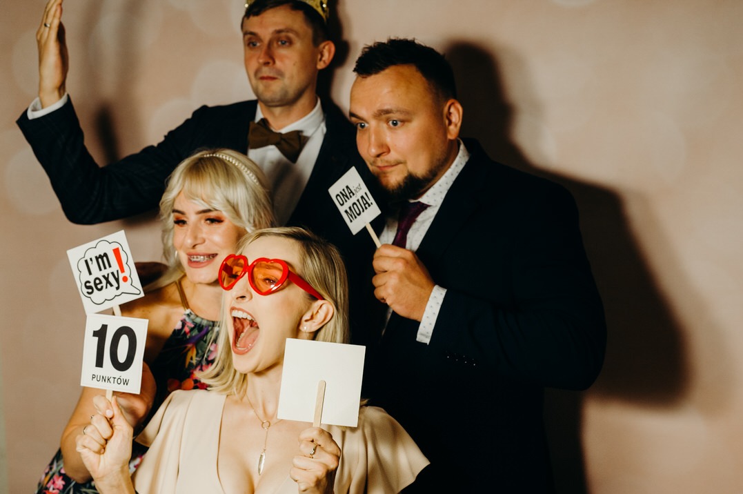 Grupa czterech osób pozuje z rekwizytami w fotobudce: kobieta w okularach w kształcie serc krzyczy, mężczyzna w koronie unosi rękę, a pozostali trzymają tabliczki z napisami.