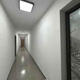 Długi korytarz w budynku, świeżo po malowaniu na biało, z widocznymi wiaderkami po farbie w rogu i oknem z widokiem na schody oraz prowizoryczne zabezpieczenia budowlane.