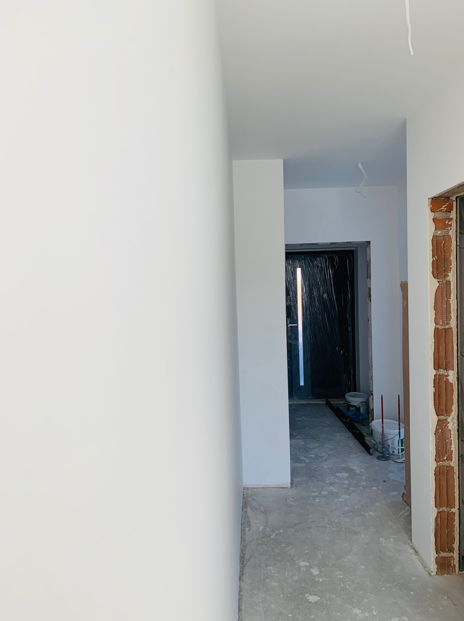 Korytarz w trakcie prac wykończeniowych, ściany pokryte świeżą, białą warstwą, zabezpieczone drzwi wejściowe folią, widoczne narzędzia i pojemniki malarskie.