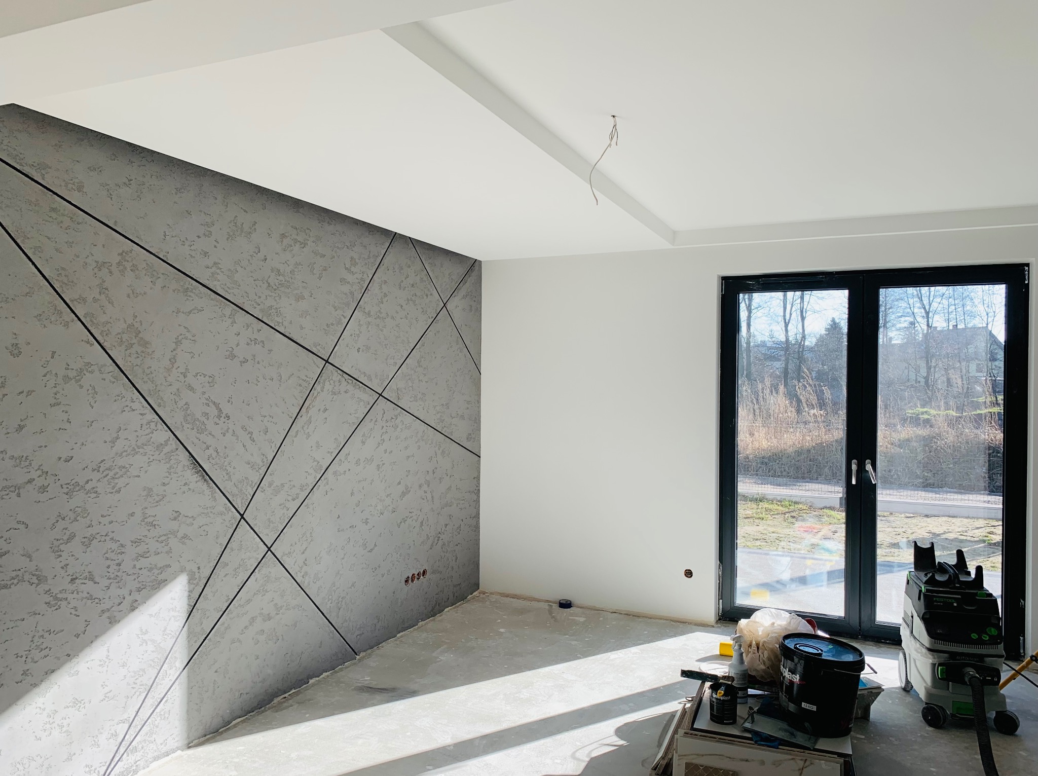 Pokój w trakcie prac wykończeniowych: ściana z dekoracyjną szarą strukturą z geometrycznym wzorem, białe ściany, czarne okno balkonowe, narzędzia i odkurzacz przemysłowy na podłodze.