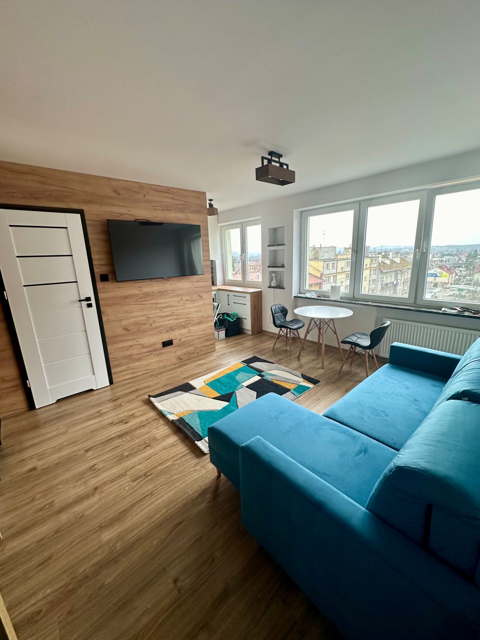 Nowoczesne wnętrze po remoncie: błękitny narożnik, drewniana ściana z telewizorem, białe drzwi z czarnymi akcentami, parkiet i dywan w geometryczne wzory, widok na miasto z okna.