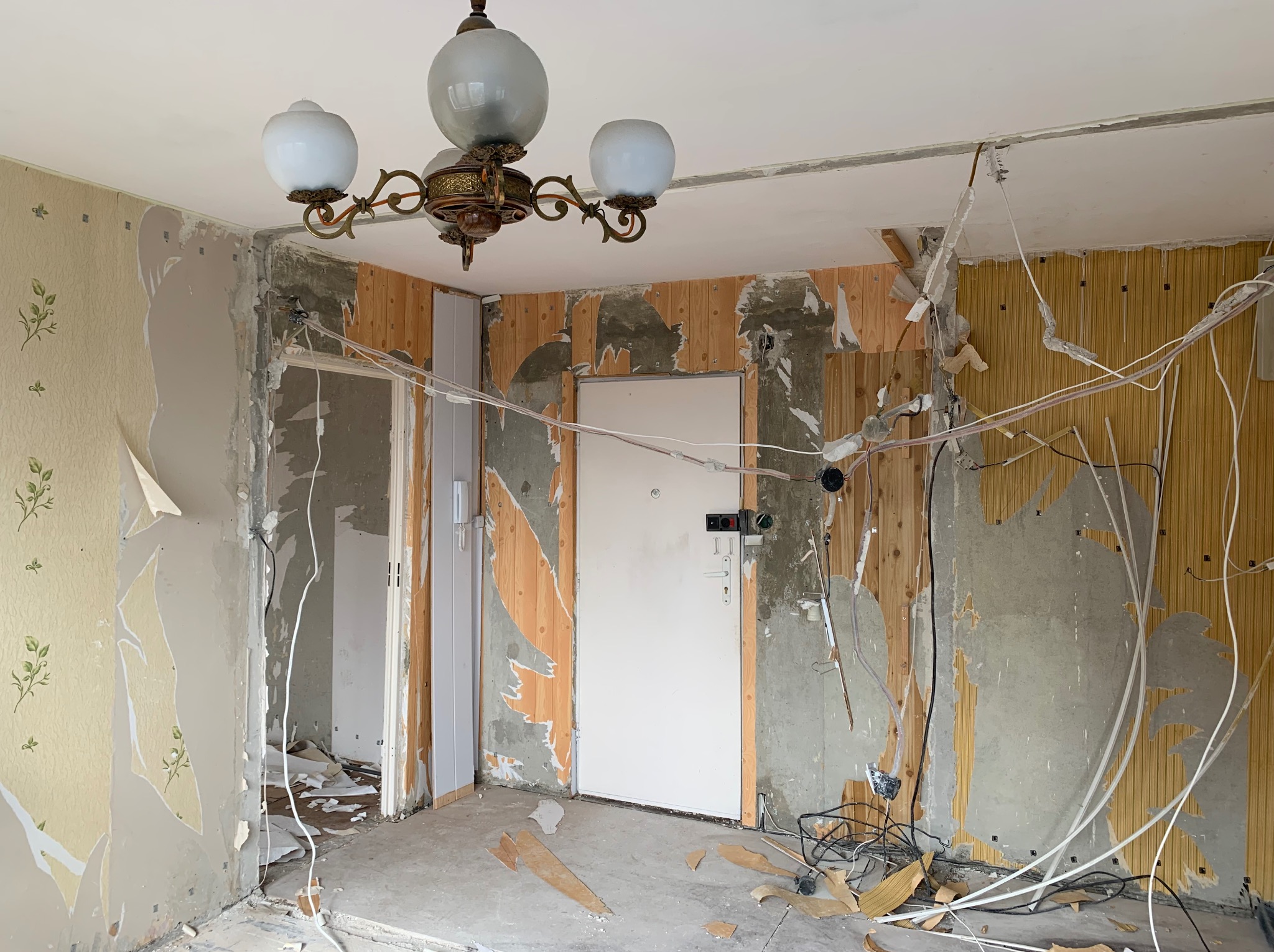 Wnętrze w trakcie demontażu: zerwane tapety z motywem roślinnym, odsłonięte drewniane panele ścienne, zwisające przewody elektryczne i centralnie umieszczona biała lampa sufitowa z trzema kloszami.