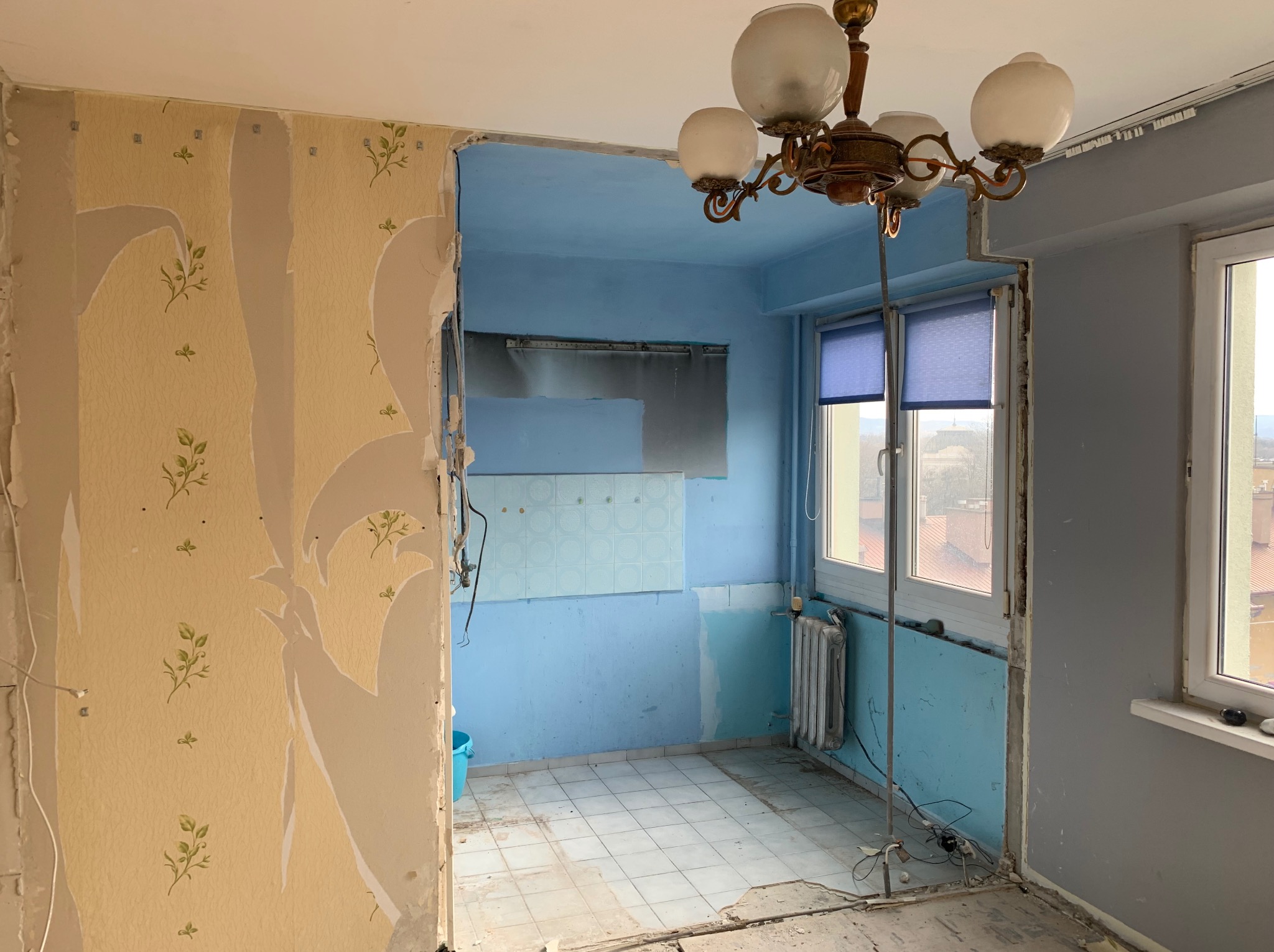 Wnętrze mieszkania w trakcie remontu: częściowo zerwana tapeta w kwiaty, pomalowane na niebiesko ściany w kolejnym pomieszczeniu, widoczny grzejnik i fragment okna.