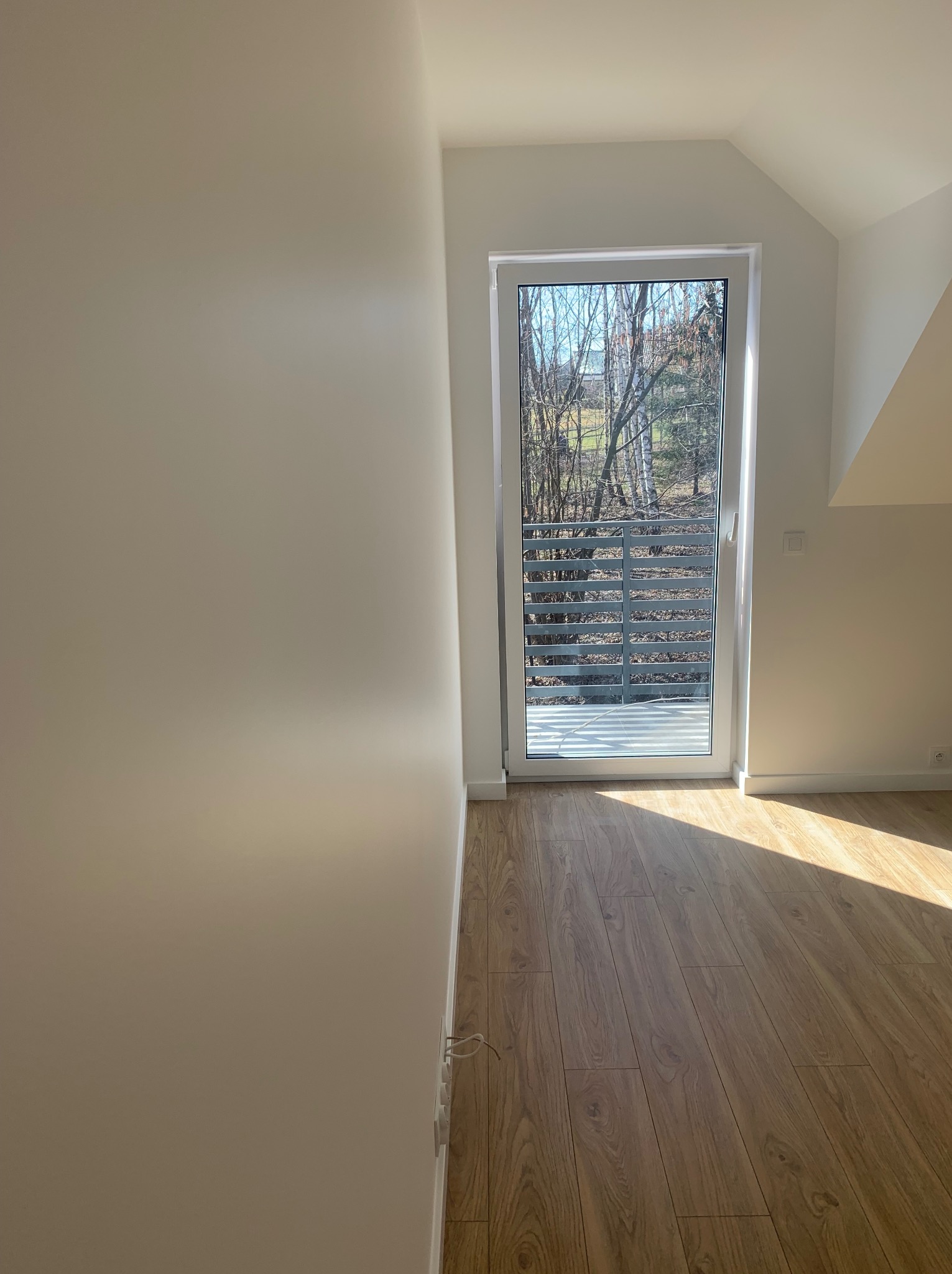 Jasne wnętrze poddasza z panelami podłogowymi w odcieniach drewna, świeżo pomalowane ściany w kolorze złamanej bieli i balkon z widokiem na drzewa bez liści w słoneczny dzień.