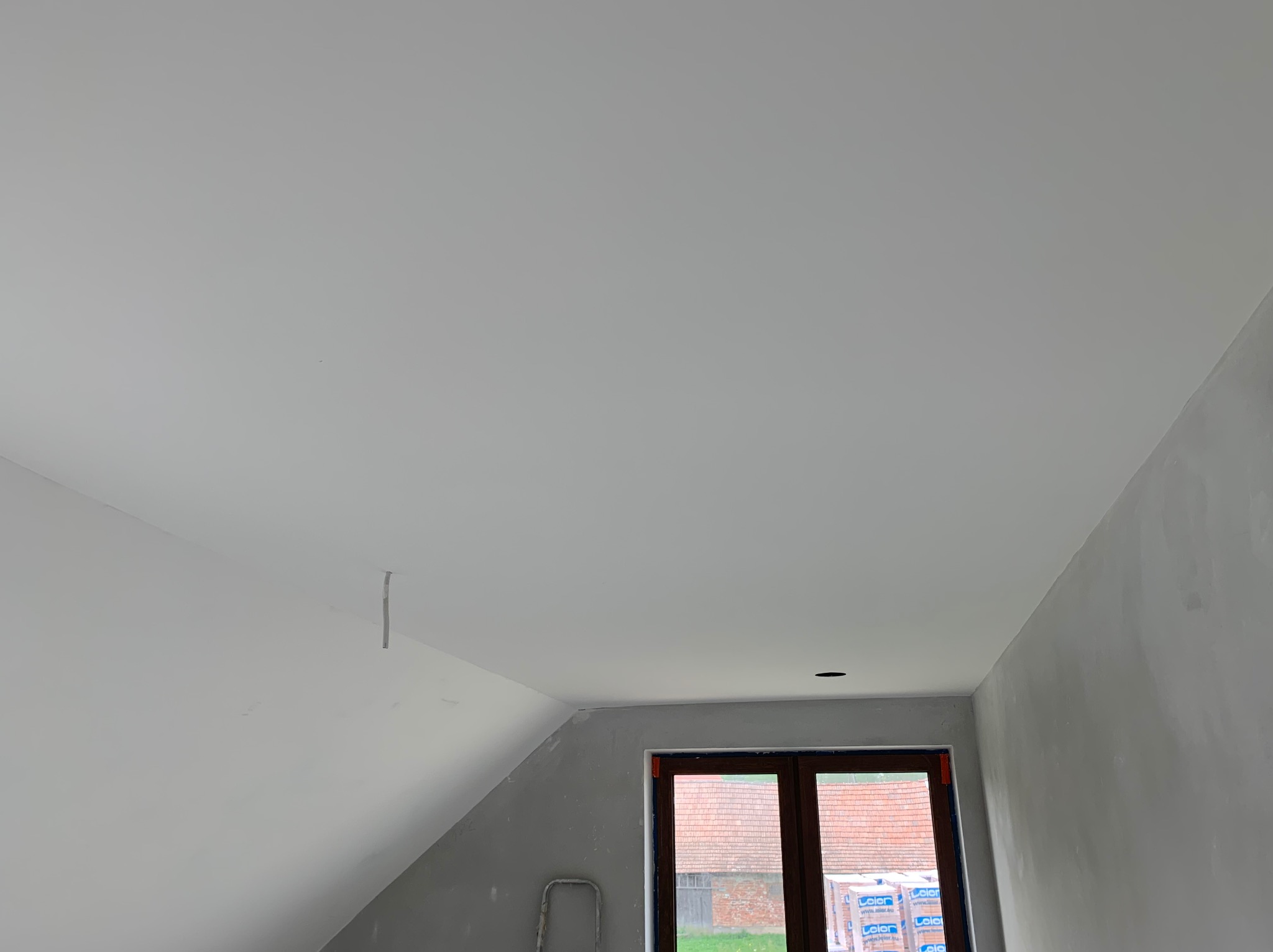 Poddasze w trakcie prac wykończeniowych: gładka, biała powierzchnia sufitu przechodzi w szpachlowaną, szarą ścianę. Widoczne okno z brązowymi ramami i fragment drabiny.