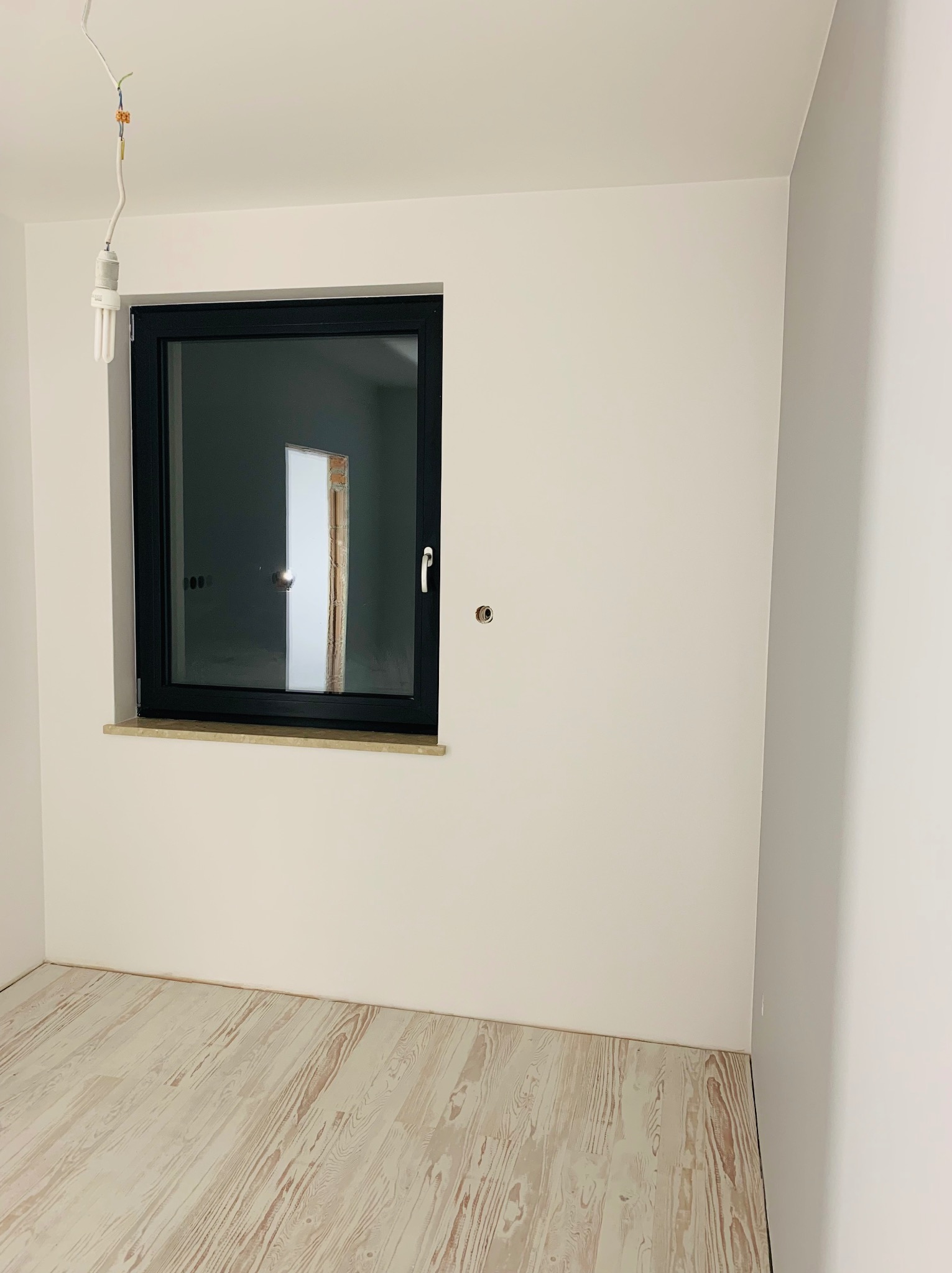 Pusty pokój po remoncie z białymi ścianami, świeżo położonymi panelami podłogowymi i czarnym oknem; widoczna wisząca żarówka na kablu.