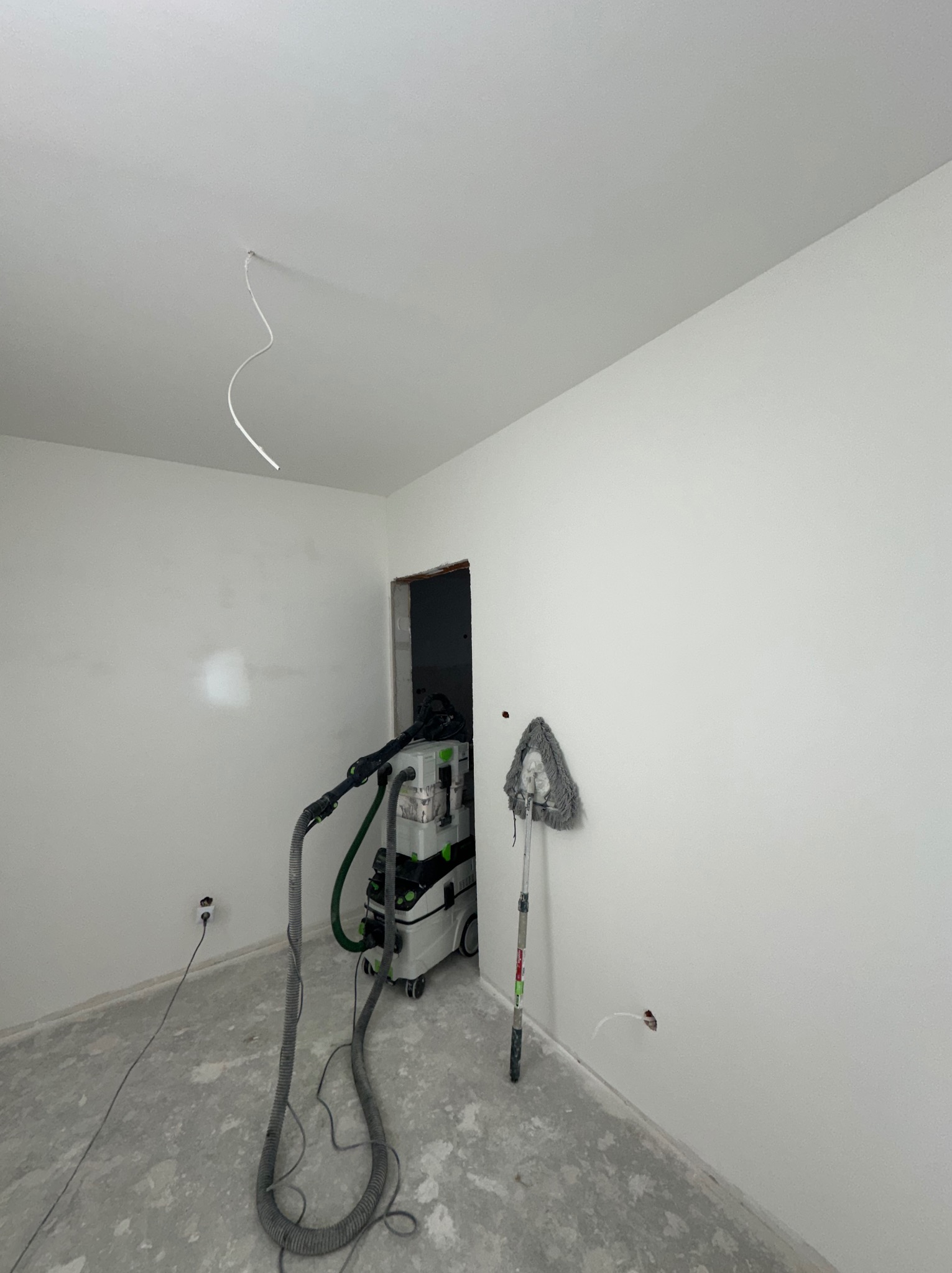 Pokój w trakcie prac wykończeniowych: gładkie, białe ściany, szlifierka do gipsu z odkurzaczem przemysłowym Festool, kabel zwisający z sufitu, widoczne ślady po szpachlowaniu.