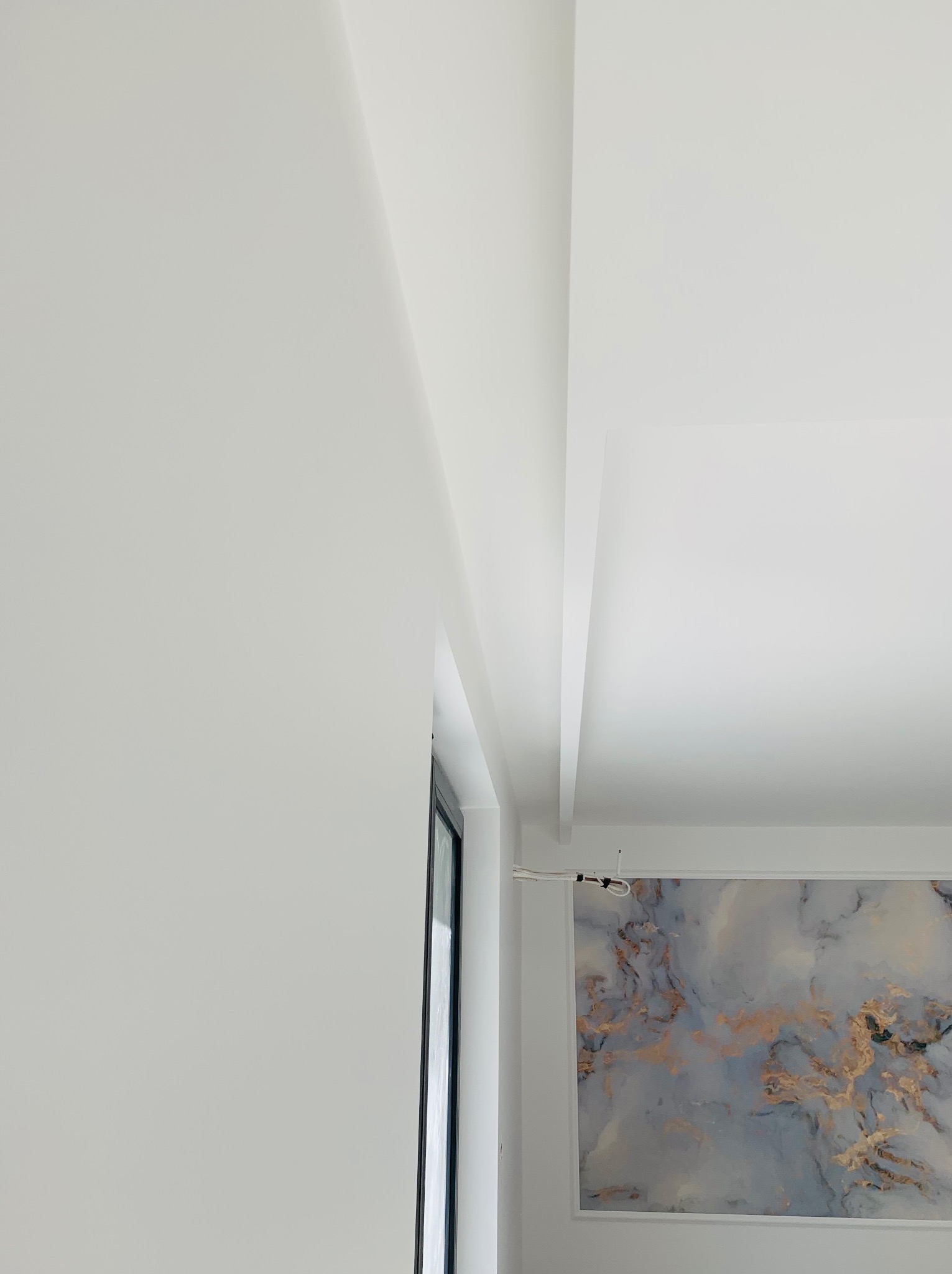 Biała ściana i sufit z dekoracyjnym panelem ściennym imitującym marmur w odcieniach szarości i złota, widoczne okno z czarną ramą.