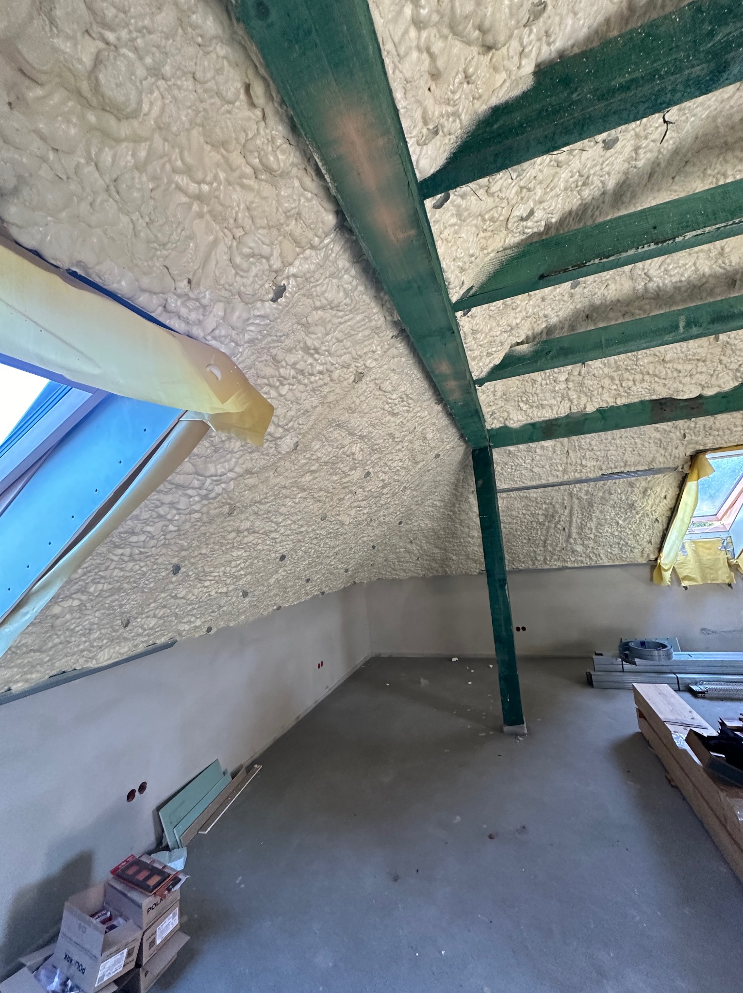 Poddasze w trakcie ocieplania pianą poliuretanową, widoczne zielone belki konstrukcyjne, okno dachowe zabezpieczone folią, surowy beton na ścianach i podłodze.