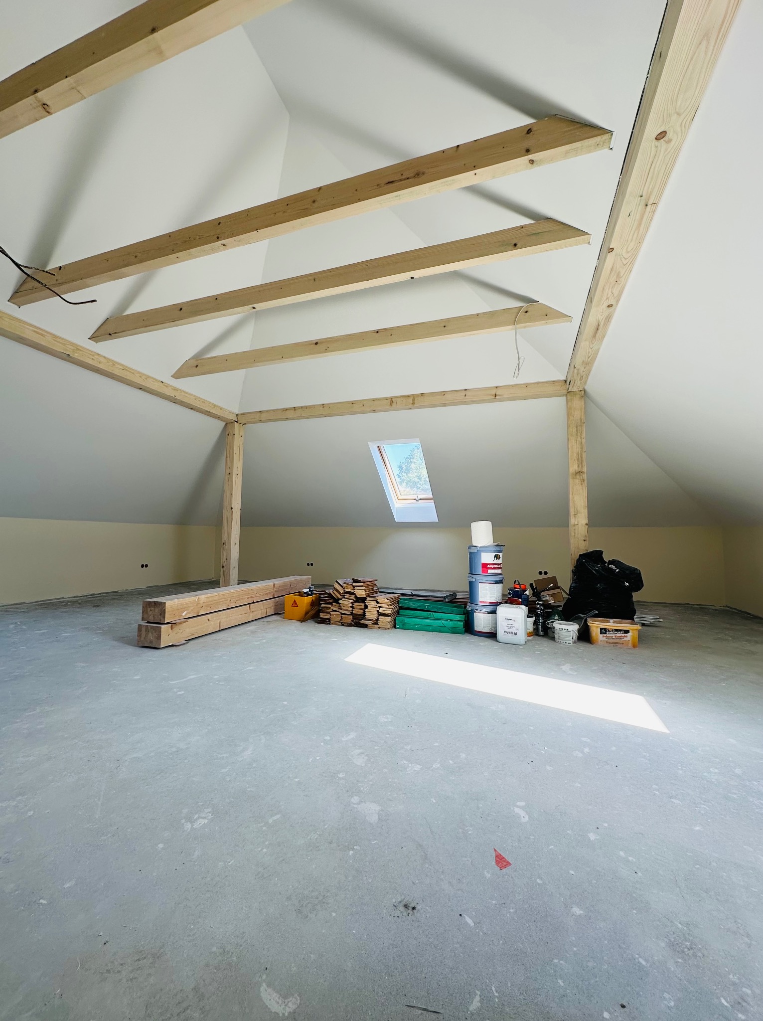 Poddasze w trakcie adaptacji: widoczne drewniane elementy konstrukcyjne, okno dachowe, przygotowane materiały budowlane i farby.