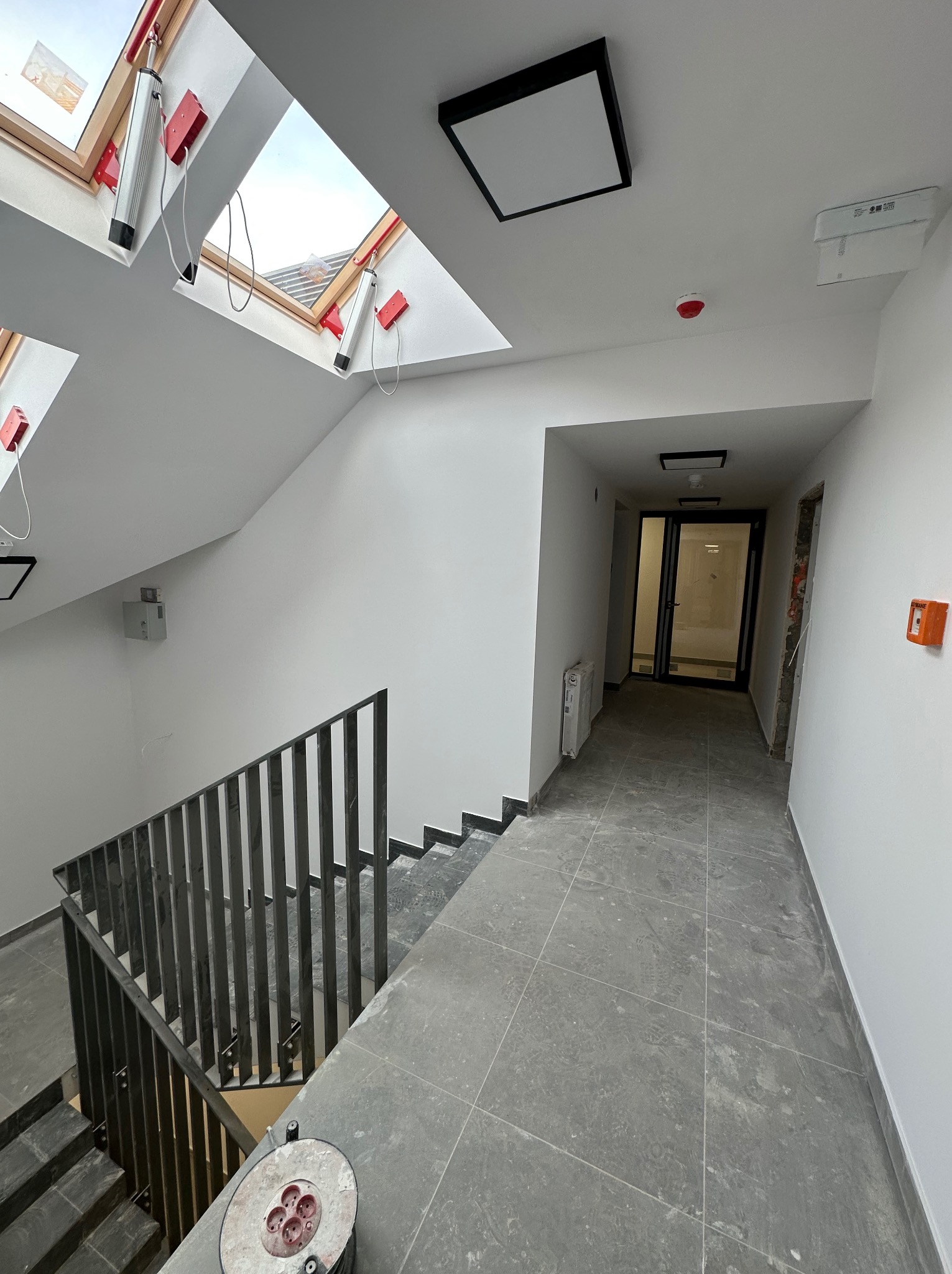 Klatka schodowa w trakcie prac wykończeniowych, widoczne gładkie, pomalowane na biało ściany, szare płytki na podłodze i schodach, czarna metalowa balustrada, okna dachowe z roletami oraz zwinięty...