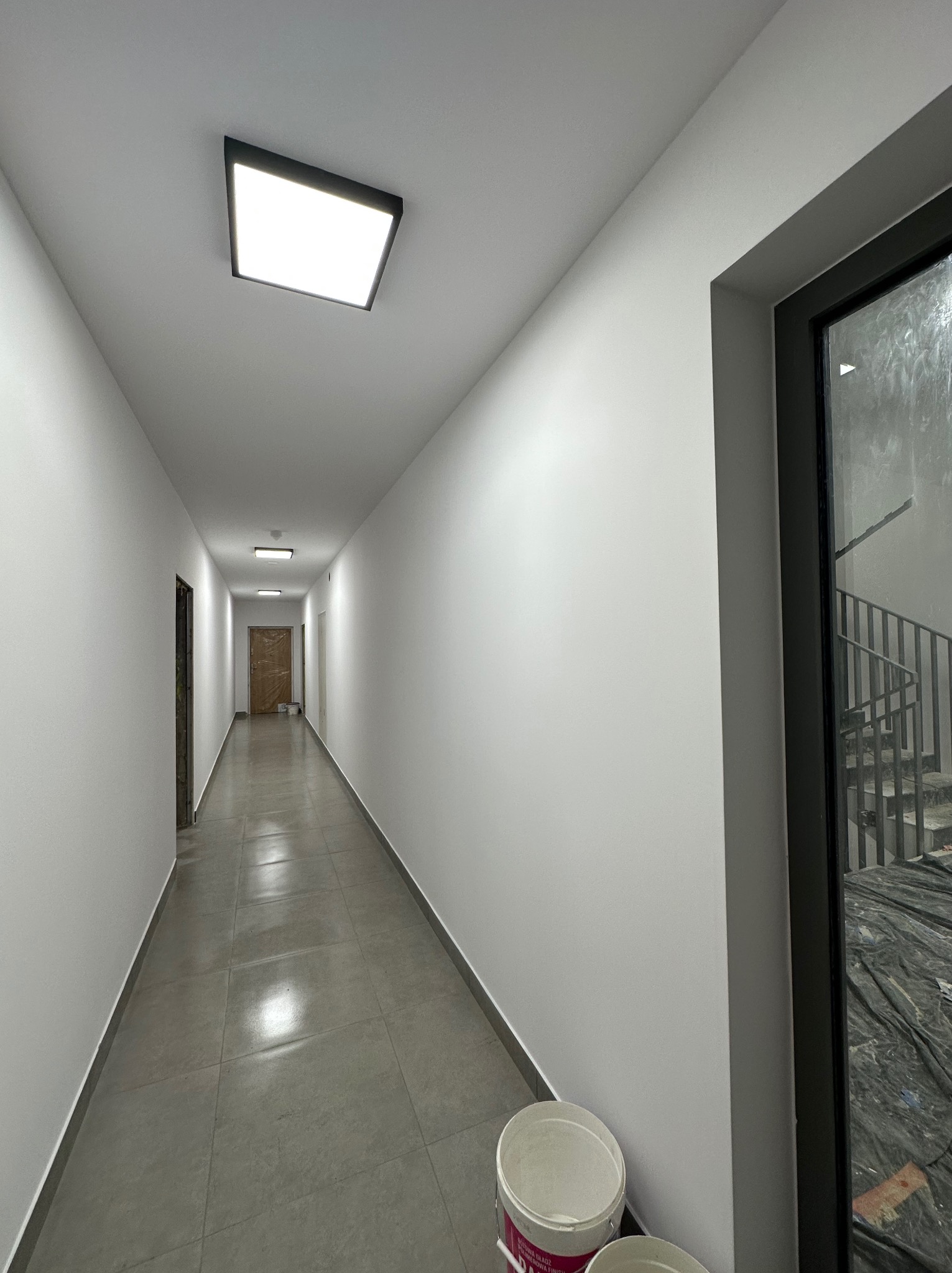 Długi korytarz w budynku, świeżo po malowaniu na biało, z widocznymi wiaderkami po farbie w rogu i oknem z widokiem na schody oraz prowizoryczne zabezpieczenia budowlane.