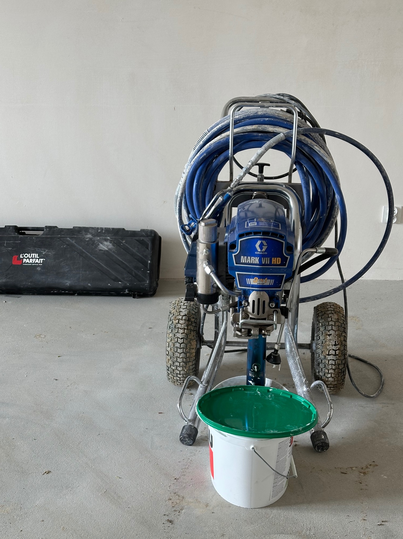Agregat malarski Graco Mark VII HD z niebieskim wężem i wiaderkiem z zieloną pokrywką na tle surowej ściany i czarnej skrzynki L'Outil Parfait.