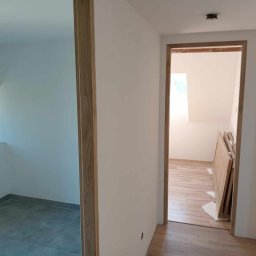 POL - DOM DANIEL NOWOTYŃSKI - Wnętrze mieszkania po remoncie: widok na dwa pomieszczenia z nowymi podłogami (szare płytki i panele imitujące drewno), świeżo pomalowane ściany na biało oraz drewniane ościeżnice drzwiowe;...