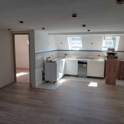 POL - DOM DANIEL NOWOTYŃSKI - Wnętrze w trakcie remontu: częściowo zamontowana biała zabudowa kuchenna, widoczne okna dachowe, podłoga wyłożona panelami, surowa ściana z przygotowanymi punktami świetlnymi.