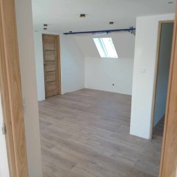 POL - DOM DANIEL NOWOTYŃSKI - Wykończone poddasze z drewnianą podłogą, oknem dachowym i dwiema drewnianymi ościeżnicami, widok z korytarza, zabezpieczone elementy sufitu taśmą malarską.