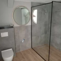 Nowoczesna łazienka z szarą glazurą, drewnopodobną podłogą, okrągłym lustrem z podświetleniem LED i kabiną prysznicową typu walk-in z czarnymi ramami.