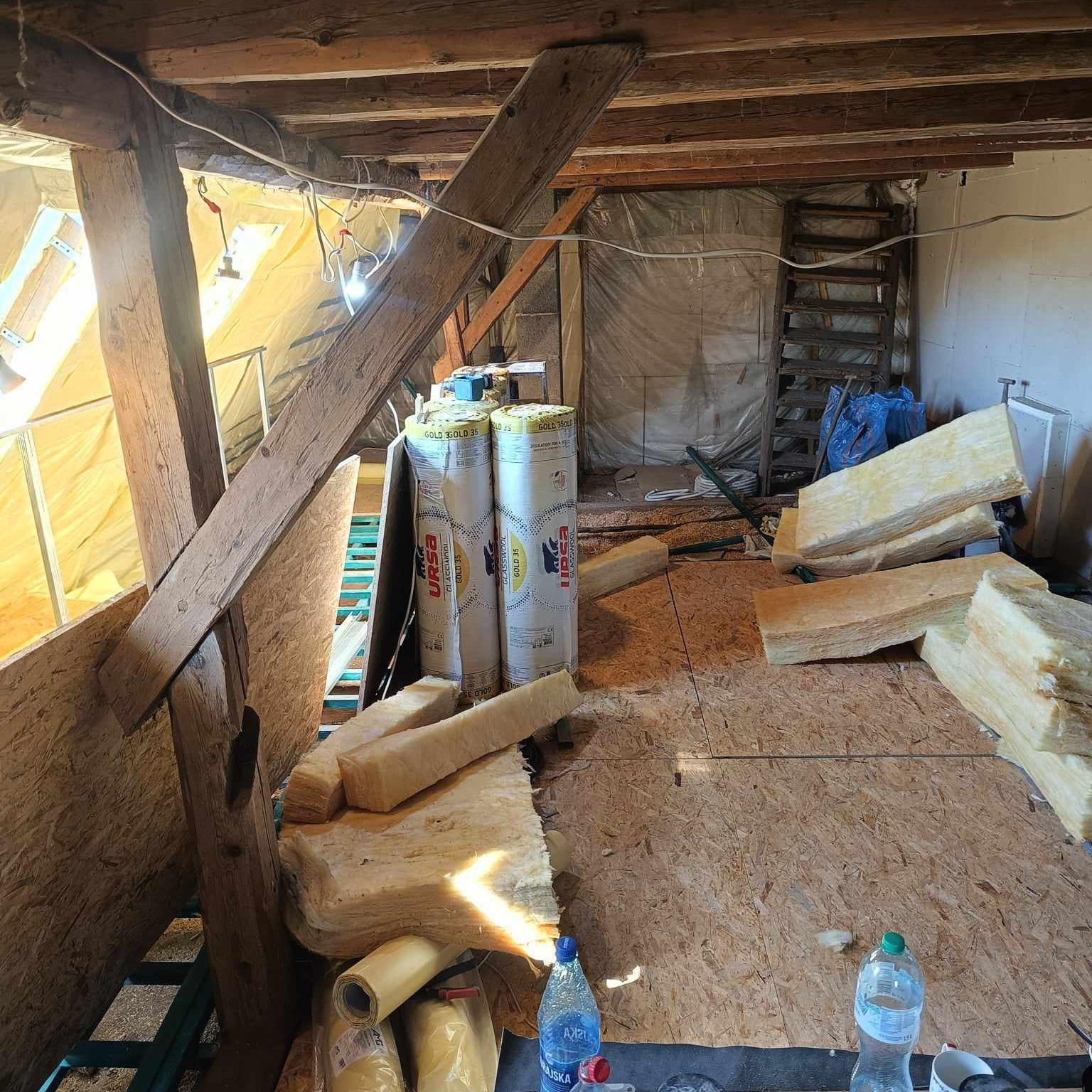 Poddasze w trakcie ocieplania, widoczne drewniane belki stropowe, płyty OSB na podłodze, rolki i fragmenty wełny mineralnej URSA, butelki wody, drabina w tle.