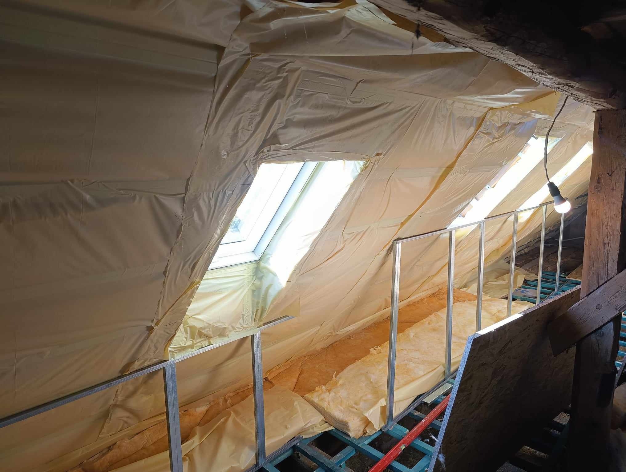 Izolacja poddasza wełną mineralną i folią paroizolacyjną w Kudowie-Zdroju, widoczne okno dachowe i metalowy stelaż.