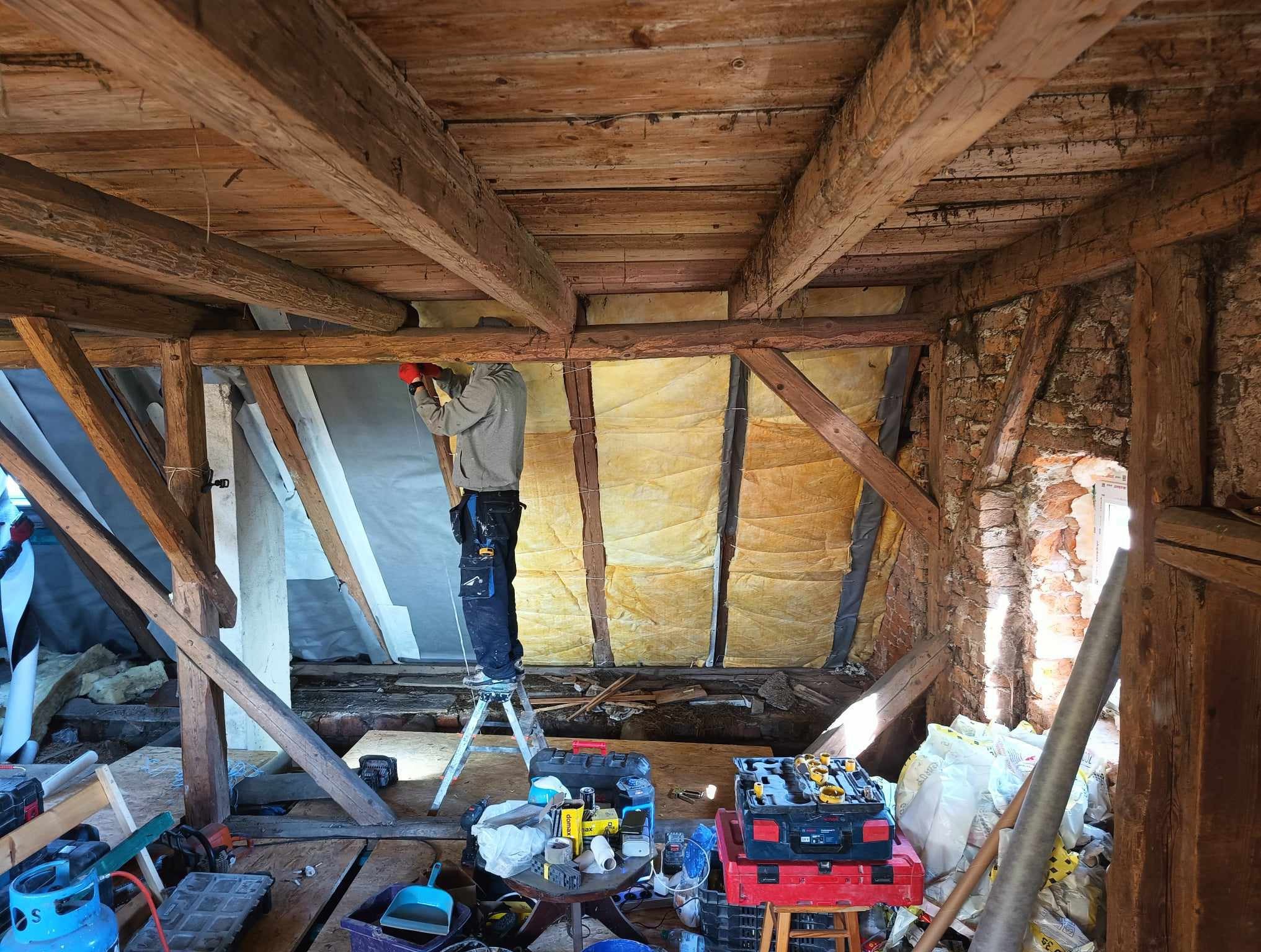 Pracownik w czerwonych rękawicach na drabinie montuje żółtą izolację termiczną między drewnianymi belkami stropu na poddaszu, widoczne narzędzia i skrzynki narzędziowe.
