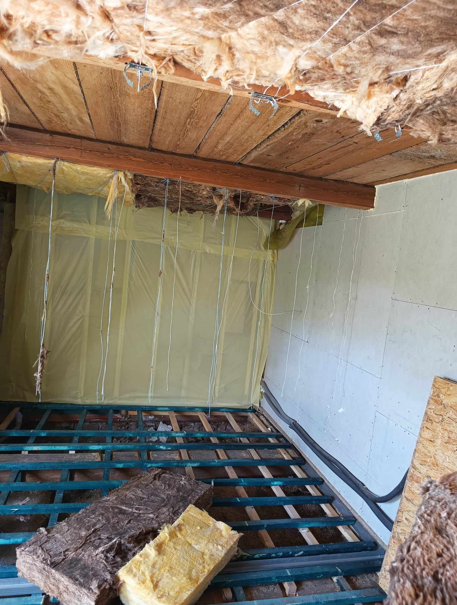 Wnętrze w trakcie remontu: izolacja wełną mineralną na poddaszu, widoczne drewniane krokwie z metalowymi uchwytami, folia paroizolacyjna na ścianie, podłoga z legarami, fragment płyty OSB.