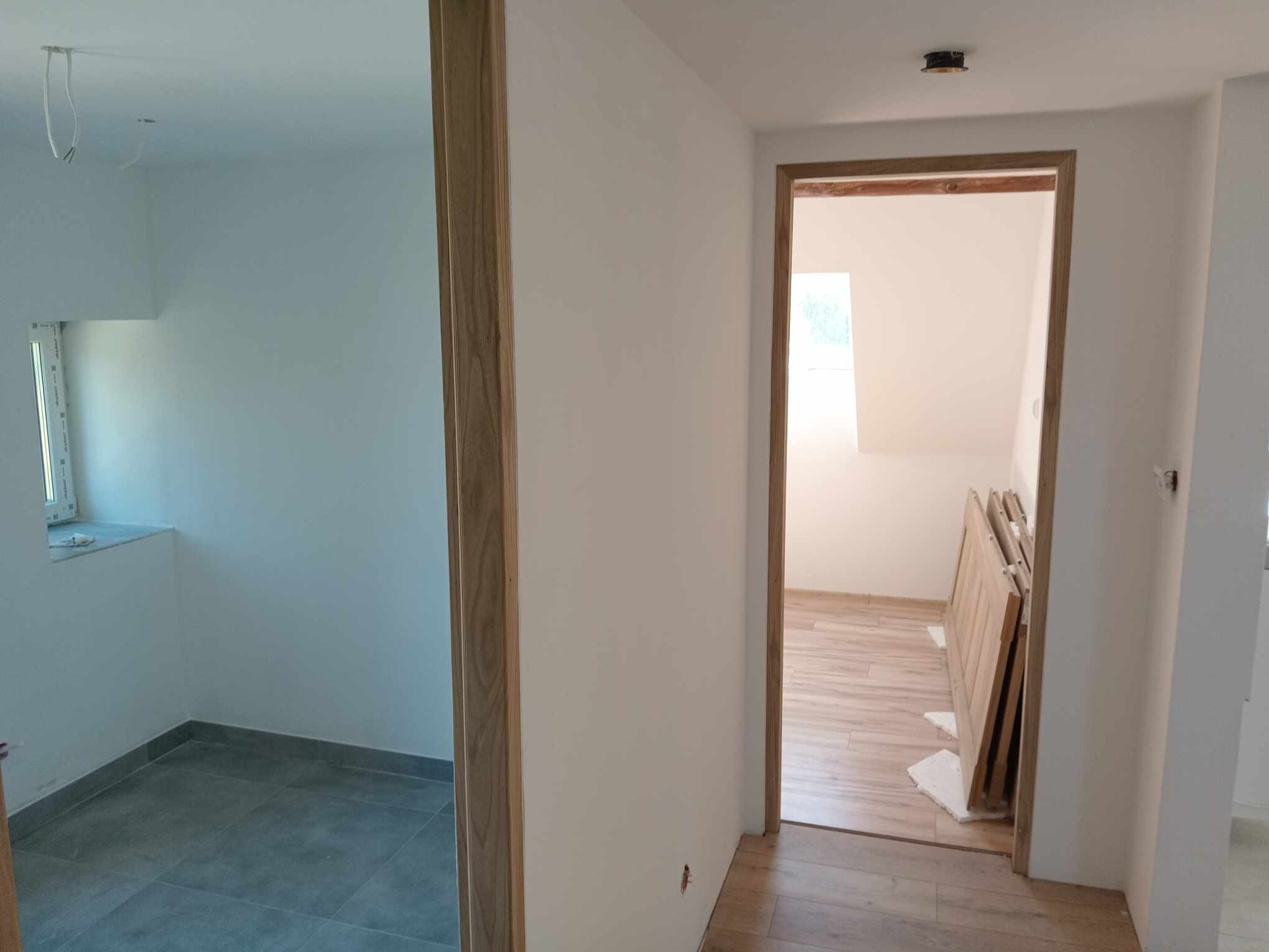 Wnętrze mieszkania po remoncie: widok na dwa pomieszczenia z nowymi podłogami (szare płytki i panele imitujące drewno), świeżo pomalowane ściany na biało oraz drewniane ościeżnice drzwiowe;...