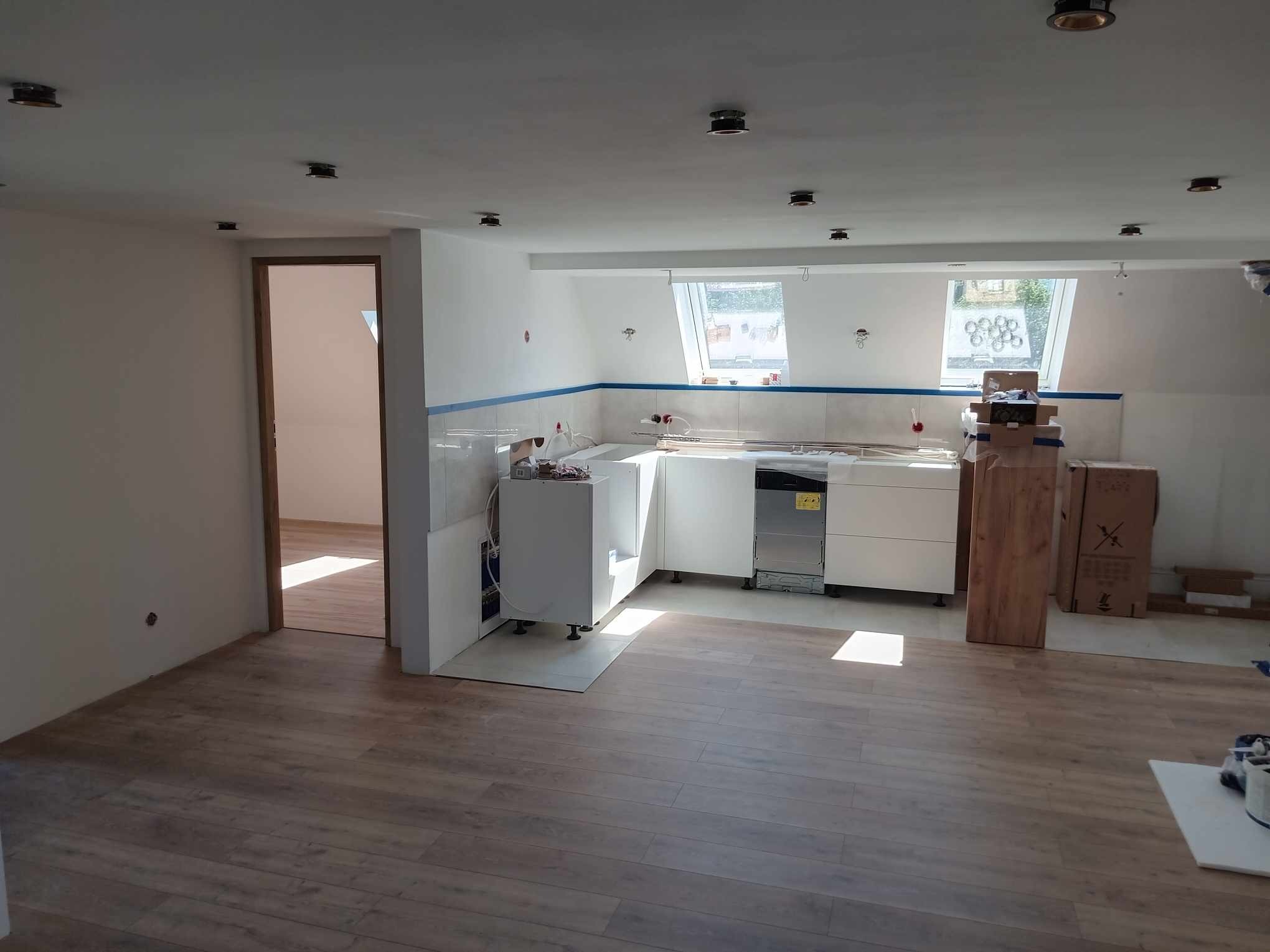 Wnętrze w trakcie remontu: częściowo zamontowana biała zabudowa kuchenna, widoczne okna dachowe, podłoga wyłożona panelami, surowa ściana z przygotowanymi punktami świetlnymi.