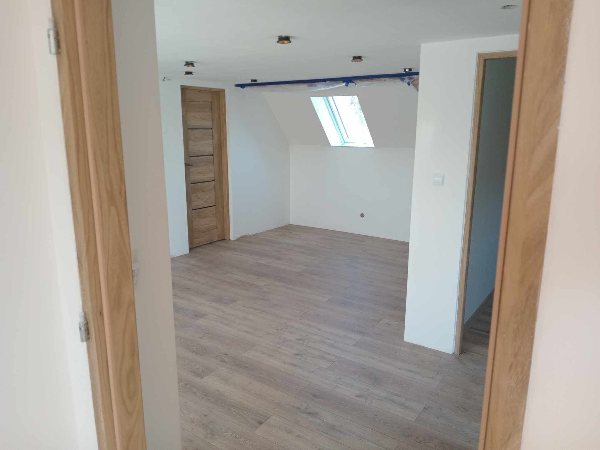 Wykończone poddasze z drewnianą podłogą, oknem dachowym i dwiema drewnianymi ościeżnicami, widok z korytarza, zabezpieczone elementy sufitu taśmą malarską.