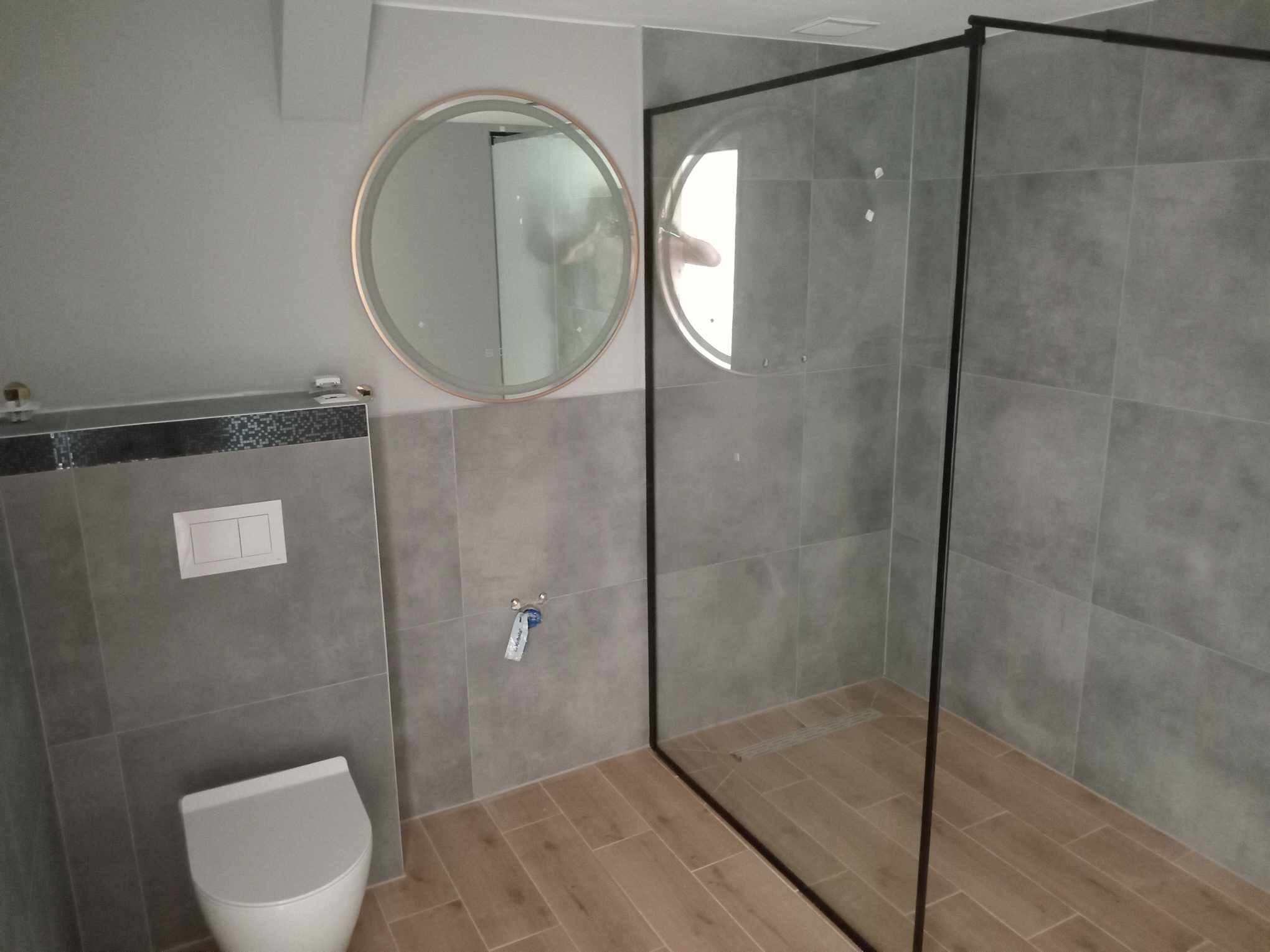 Nowoczesna łazienka z szarą glazurą, drewnopodobną podłogą, okrągłym lustrem z podświetleniem LED i kabiną prysznicową typu walk-in z czarnymi ramami.