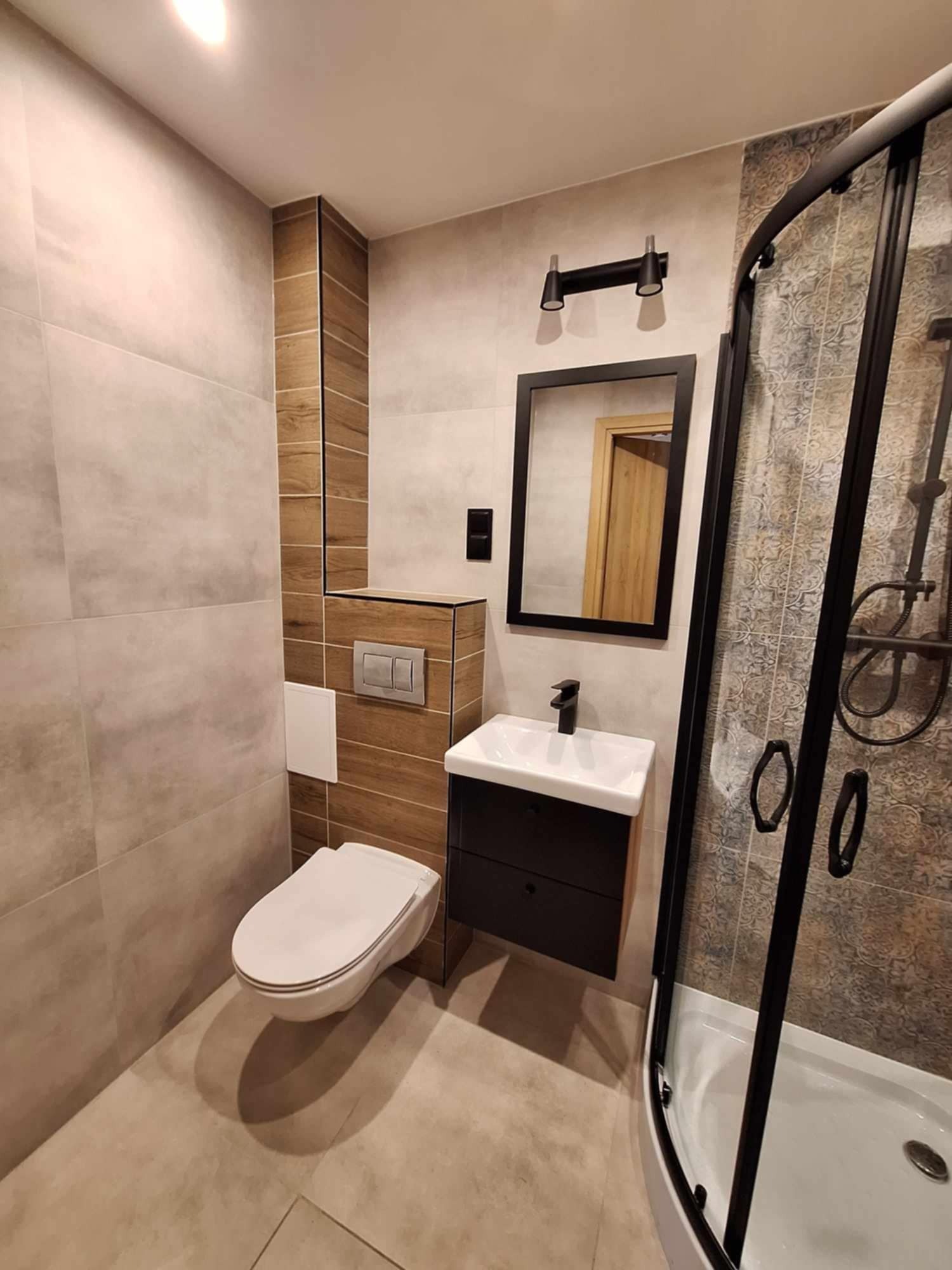 Wykończona łazienka z beżowymi płytkami imitującymi kamień, drewnopodobną obudową geberitu, czarną kabiną prysznicową z wzorzystymi płytkami wewnątrz, czarną szafką pod umywalką i lustrem w czarnej...