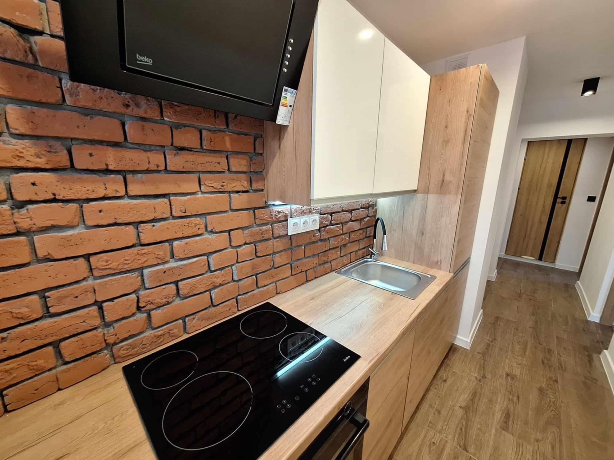 Nowoczesna kuchnia z ceglaną ścianą, drewnianymi szafkami i czarną płytą indukcyjną, widok na korytarz z drewnianymi drzwiami.