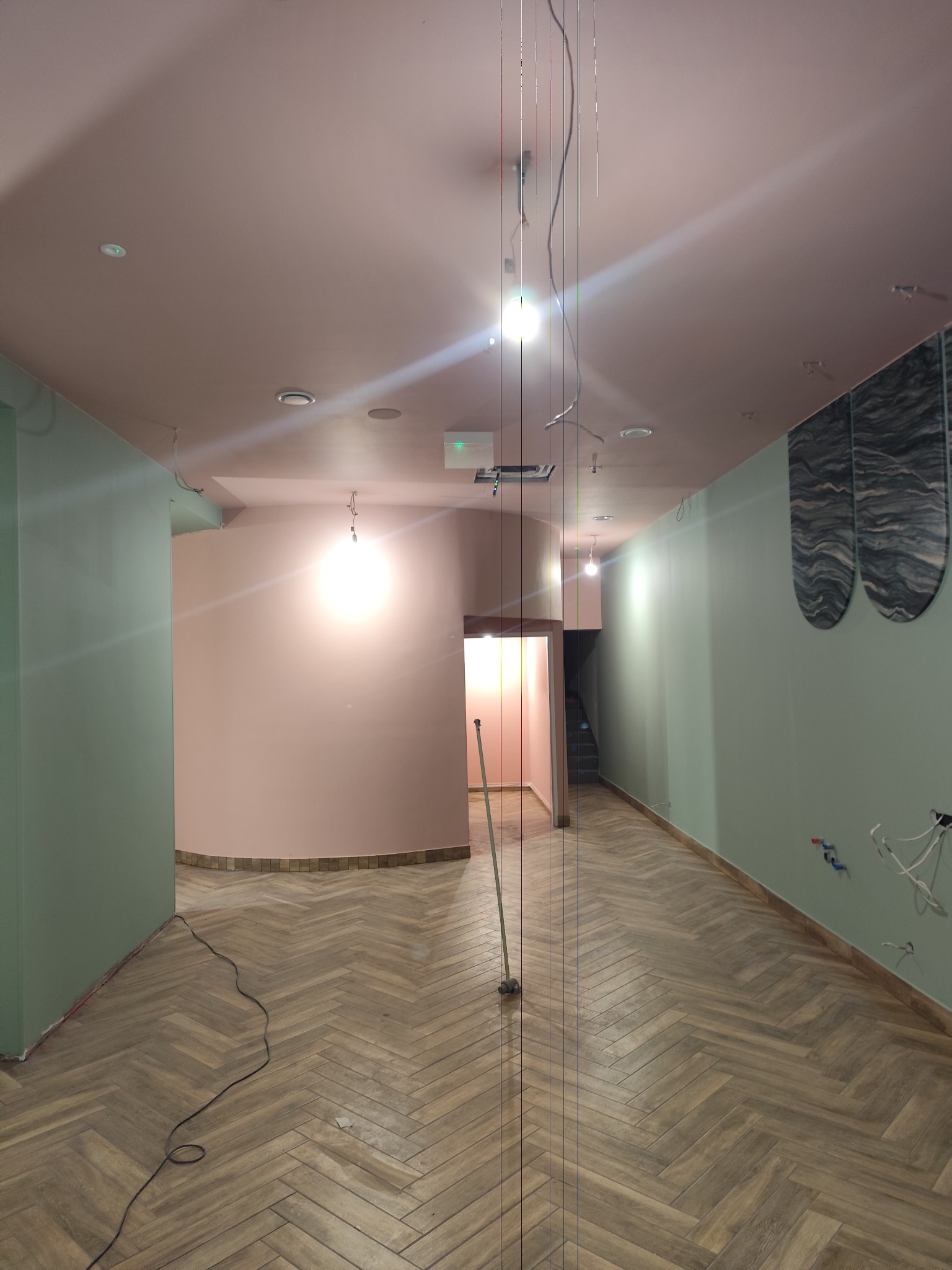 Wnętrze w trakcie prac wykończeniowych: parkiet w jodełkę, ściany w odcieniach zieleni i różu, widoczne przewody i narzędzia, sufit z oświetleniem.