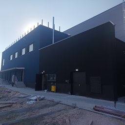 REMWIK Joanna Pęczkowska - Nowoczesny budynek przemysłowy z niebieską i czarną elewacją, widoczne elementy konstrukcyjne i teren wokół w trakcie prac wykończeniowych, z grupką osób w odblaskowych kamizelkach w tle.