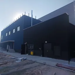 Nowoczesny budynek przemysłowy z niebieską i czarną elewacją, widoczne elementy konstrukcyjne i teren wokół w trakcie prac wykończeniowych, z grupką osób w odblaskowych kamizelkach w tle.
