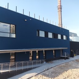 REMWIK Joanna Pęczkowska - Niebieski budynek przemysłowy z widocznym kominem w tle, teren wokół w trakcie zagospodarowywania, świeżo położony chodnik z kostki.