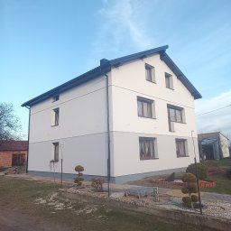 REMWIK Joanna Pęczkowska - Dwukondygnacyjny dom z nową, dwukolorową elewacją w odcieniach szarości, z ciemnym dachem i brązowymi oknami, otoczony ogrodem z ozdobnymi krzewami.