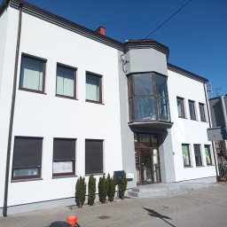 REMWIK Joanna Pęczkowska - Biały budynek z szarymi akcentami i brązowymi ramami okiennymi, z przeszklonym wykuszem nad wejściem. Widoczne rolety zewnętrzne i małe drzewka iglaste.