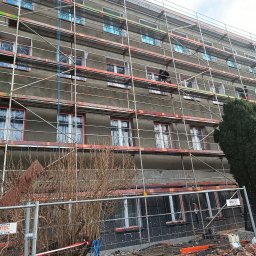 REMWIK Joanna Pęczkowska - Budynek w trakcie prac elewacyjnych, rusztowania, okna zabezpieczone folią, widoczni pracownicy na rusztowaniu, gruz i materiały budowlane na dole.