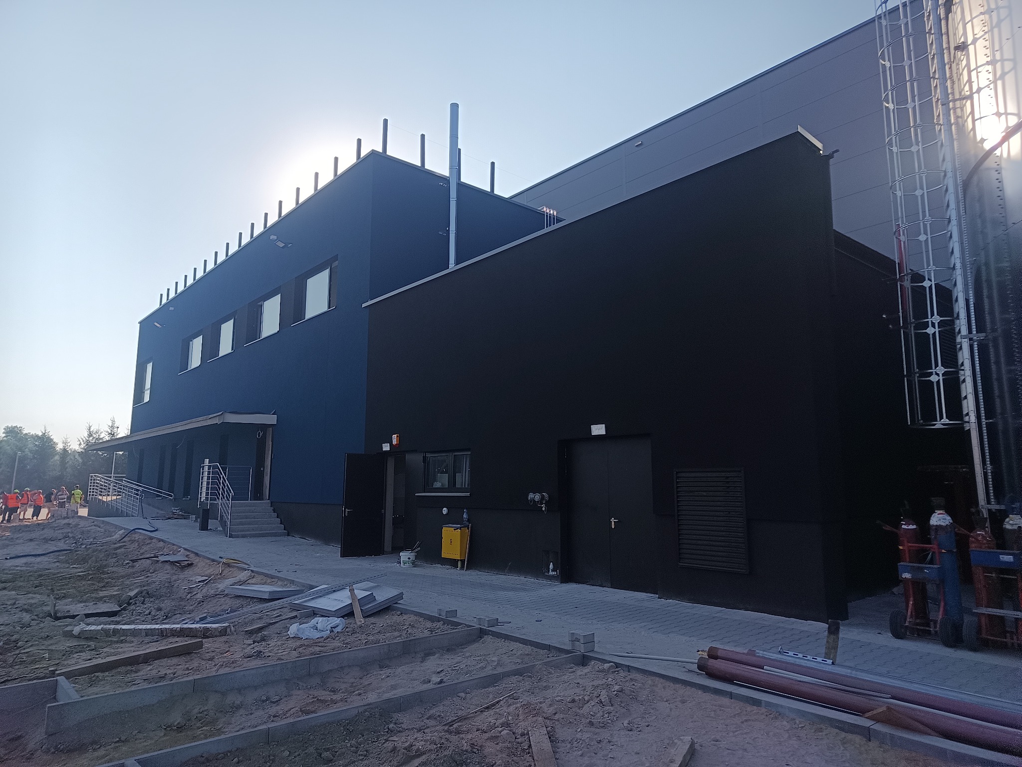 Nowoczesny budynek przemysłowy z niebieską i czarną elewacją, widoczne elementy konstrukcyjne i teren wokół w trakcie prac wykończeniowych, z grupką osób w odblaskowych kamizelkach w tle.