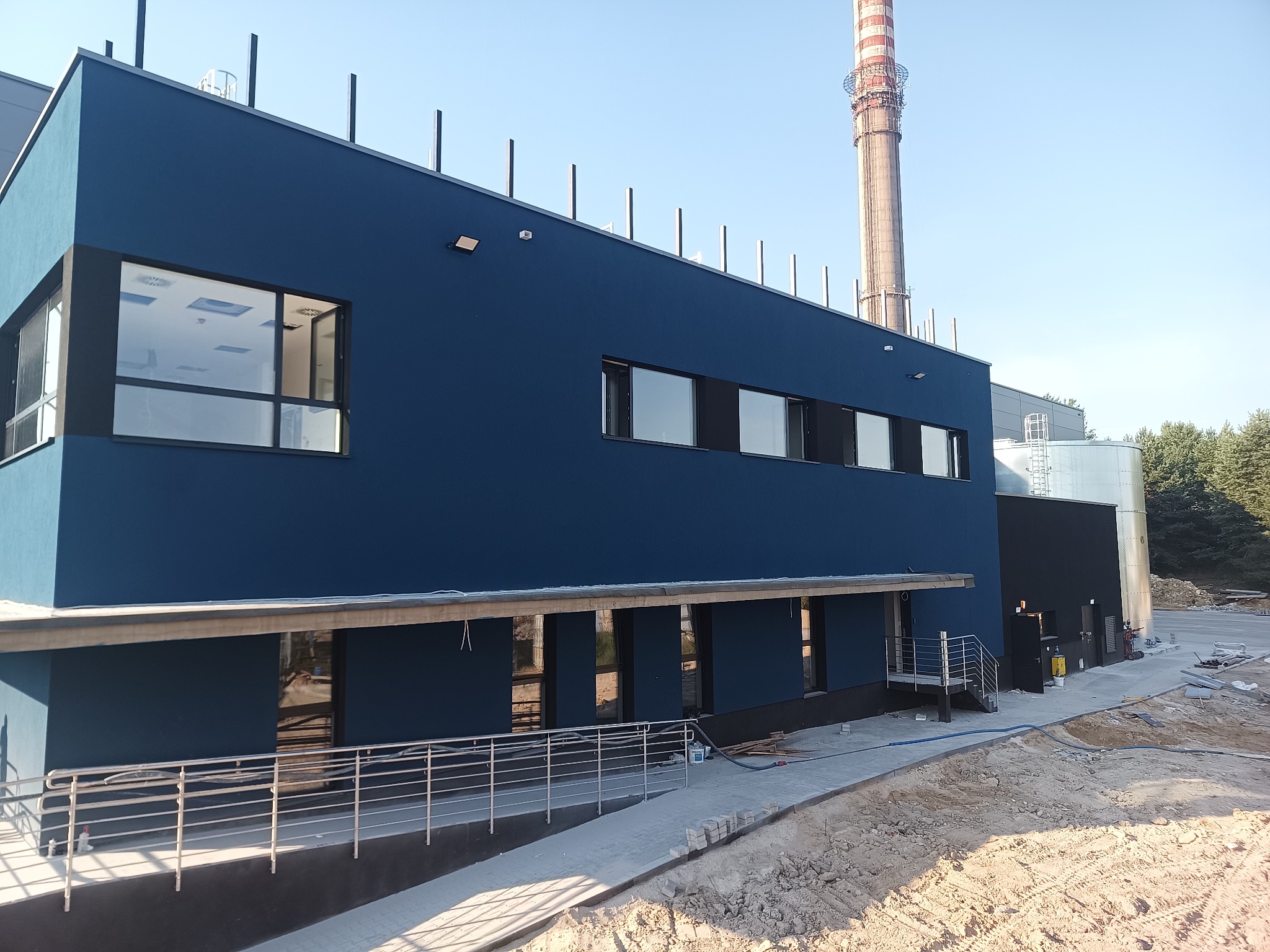 Niebieski budynek przemysłowy z widocznym kominem w tle, teren wokół w trakcie zagospodarowywania, świeżo położony chodnik z kostki.