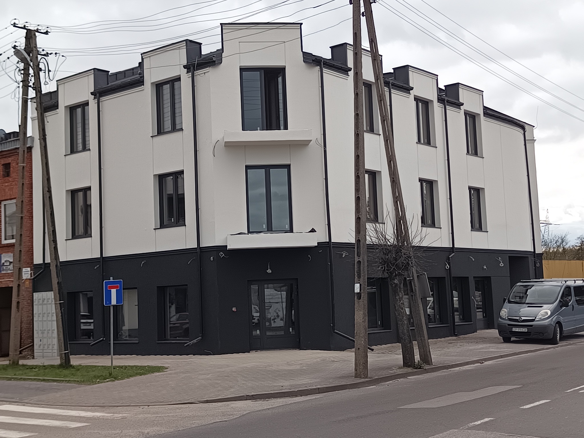 Dwukondygnacyjny budynek po remoncie elewacji z kontrastującym, ciemnym dołem i białymi ścianami, widoczny z ulicy w pochmurny dzień, z zaparkowanym szarym vanem i znakiem drogowym.