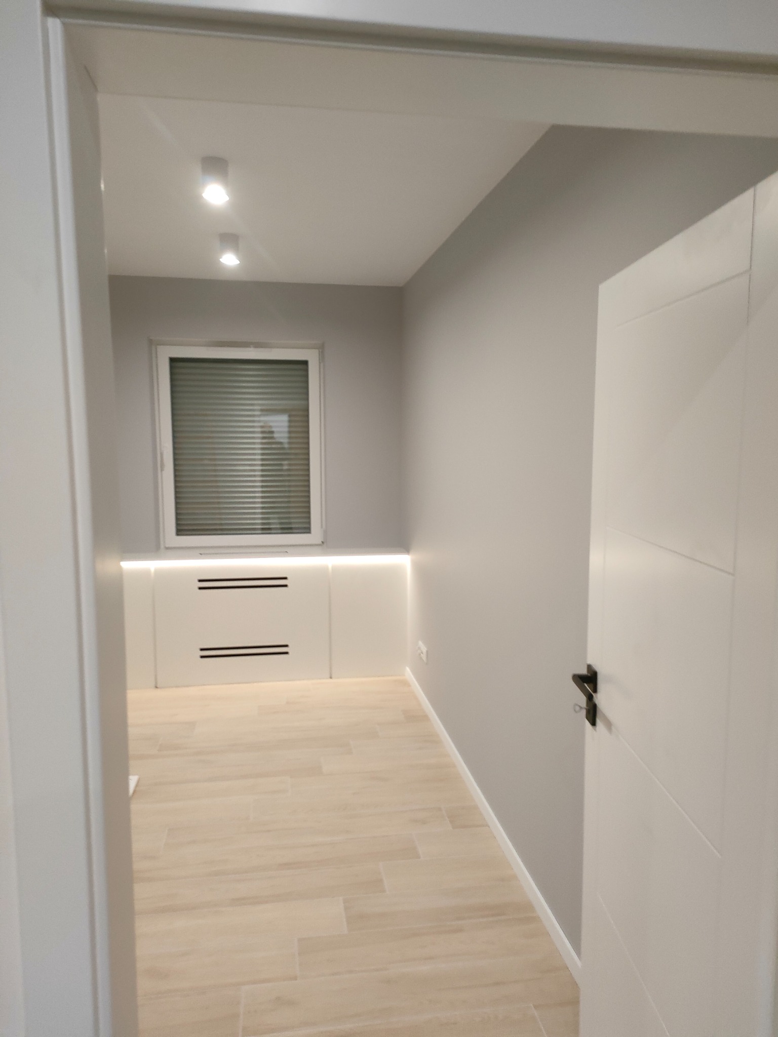 Wnętrze pokoju po remoncie: jasne ściany, kremowa podłoga z płytek imitujących drewno, białe zabudowy pod oknem z podświetleniem LED i białe drzwi z czarną klamką.