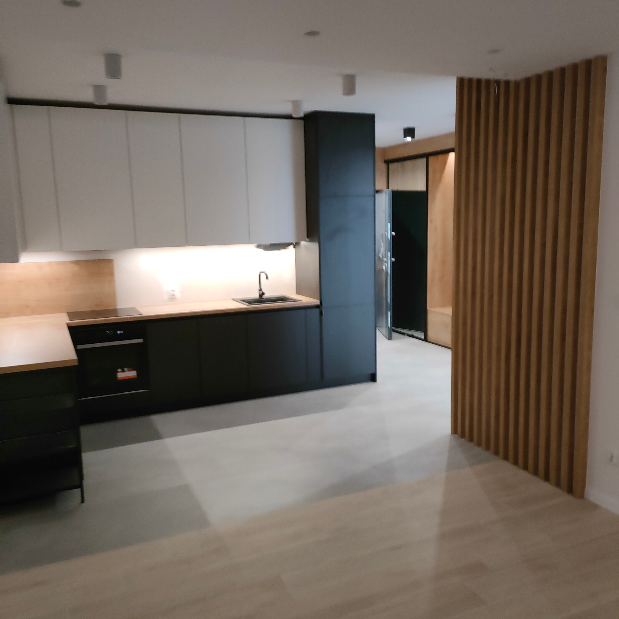 Nowoczesna kuchnia z białymi górnymi szafkami i czarnymi dolnymi, drewnianym blatem i panelem nad blatem, oraz dekoracyjną drewnianą ścianką działową z pionowych listew.